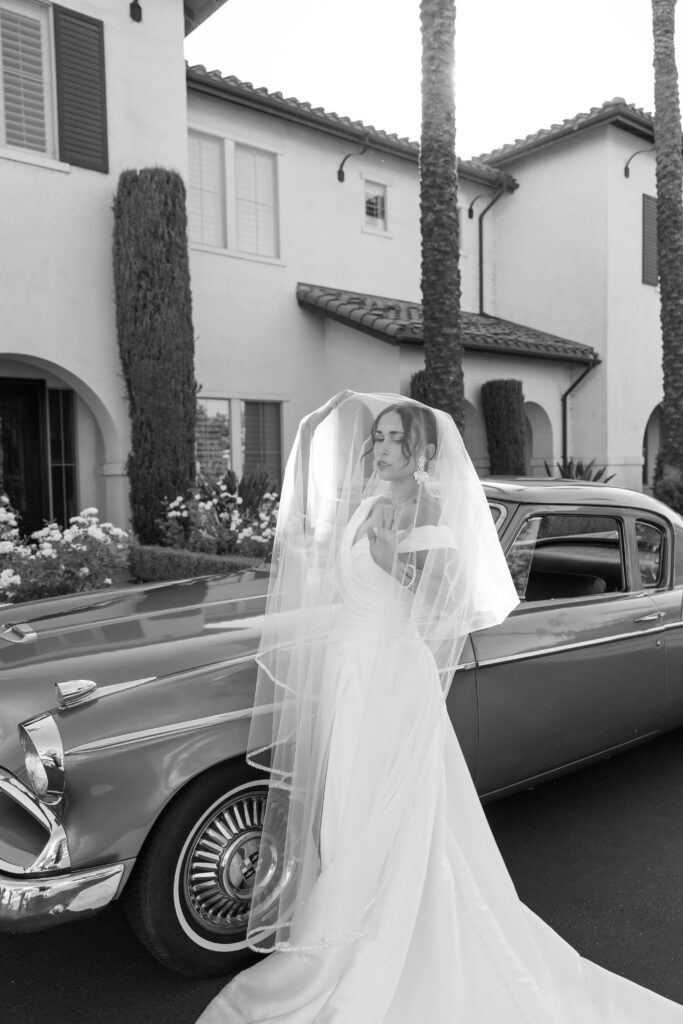 Timeless bridal portrait with flowing veil beside a vintage classic car outside a wedding venue.