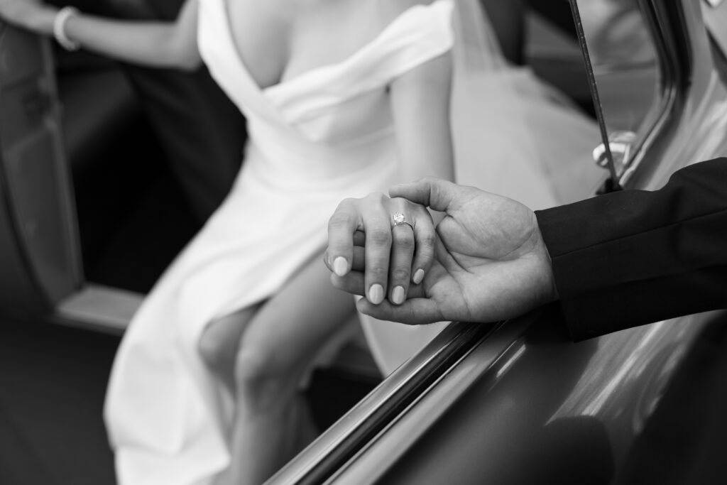 Candid black and white photo of bride and groom holding hands through the car window after their wedding ceremony.