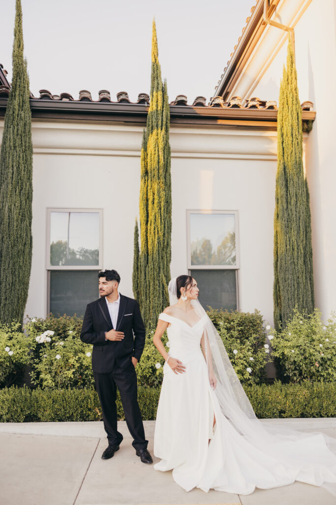 Editorial wedding portrait of a bride and groom standing together outside a Tuscan-inspired villa with tall cypress trees.